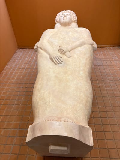 The image displays a large, light-colored anthropoid sarcophagus standing upright, viewed from a low angle, within an indoor environment. The sarcophagus, likely carved from stone or a similar material, depicts a stylized recumbent human figure. The figure has sculpted curly hair, closed eyes, a prominent nose, and a serene facial expression. Its arms are bent, with the left hand resting over the right hand across the lower torso. Bare feet with delineated toes are visible at the base of the figure, resting on a sculpted plinth. The sarcophagus stands on a wider, rectangular base with a smaller protruding block underneath. The floor is tiled with small, square, reddish-brown terracotta tiles, while the surrounding walls are painted a uniform, warm orange-brown color. This object is identified as an anthropoid sarcophagus, characteristic of Phoenician/Punic art, located in the Museo de Cádiz (Museum of Cádiz) in Cádiz, Spain.