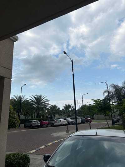 The image shows a residential street in La Aurora, Ecuador. Several cars are parked along the red brick paved road. Palm trees and other greenery line the street. Tall street lights are spaced along the road. The sky is blue with scattered clouds. The foreground includes part of a building's exterior and a car's hood. No people are visible.