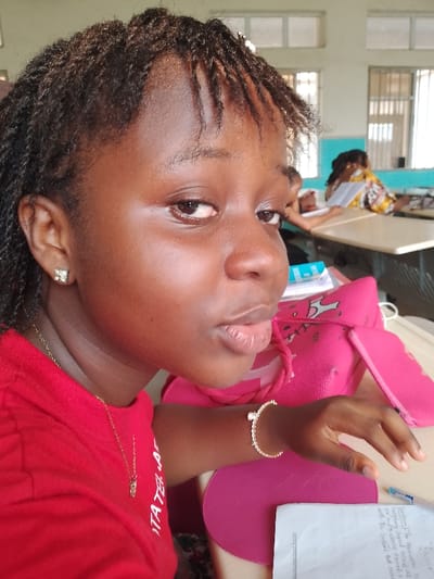 The image shows a young woman in a red shirt, wearing gold jewelry (earrings, necklace, bracelet) sitting at a desk. She is in what appears to be a classroom setting, with other students visible in the background. She is looking at the camera. There is a pink bag next to her, as well as school-related items like paper and a pen. The setting is in Ogboko, Nigeria.