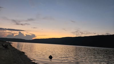 The image depicts a serene sunset over the Dnipro River in the vicinity of Kulishivka, Ukraine. Silhouetted against the twilight sky, a dark, tree-lined hill forms the far bank of the river. The water reflects the warm hues of the setting sun, with ripples breaking the smooth surface. On the sandy shore in the foreground, several individuals are casually seated, some in chairs, possibly engaged in fishing or simply enjoying the view. Fishing rods are visible along the bank, and a lone stick or piece of debris emerges from the water. The sky transitions from a pale blue at the top to a warm orange and pink glow near the horizon, with scattered clouds adding texture to the atmospheric scene. The overall impression is one of tranquility and natural beauty as day transitions into evening.