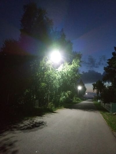 The image depicts an asphalt road in what appears to be a rural or suburban area at dusk or night. The sky is a deep blue, with some clouds visible. A bright street lamp illuminates the left side of the road, casting strong light on the surrounding trees and creating prominent shadows on the road surface. On the right side of the road, another street lamp is visible further down, and on the far right, a metal fence and grass can be seen. The overall atmosphere is dimly lit, with the artificial light from the lamps being the dominant light source. The location is identified as Voroshilovka, Kazakhstan.