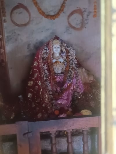 The image depicts an altar dedicated to a female deity, likely within a temple in Khakhnal, India. A marble statue of the deity, adorned with a vibrant red and gold embroidered sari, is the central focus. She wears an ornate crown and has a serene expression. Garlands of orange marigolds are draped around the altar and a red circular marking is visible on the wall behind the deity. The altar itself is constructed of wood, with decorative wooden posts supporting a shelf that displays offerings of flowers. The background wall appears to be made of rough plaster, with some areas showing signs of discoloration. The overall scene suggests a place of worship, imbued with devotional elements.