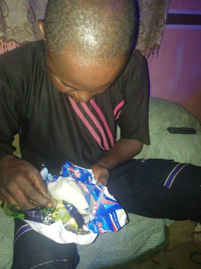 A man is sitting on a green bedspread, holding a snack bag. He is wearing a black shirt with pink stripes and black pants with red and blue stripes. The snack bag is open, revealing white contents. In the background, there are patterned curtains and a pink wall. The location is Oja Tutun, Oko Erin, Nigeria.