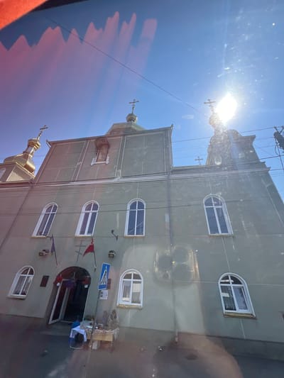 A low-angle, wide shot captures a pale green church in Kovel, Ukraine, under a bright, sunny sky. The sun is high and somewhat obscured by lens flare and atmospheric haze, casting bright rays across the scene. The church features several arched windows with white frames and a golden dome with a cross. Two flags, one purple and one red, hang from the facade near the entrance. A pedestrian crossing sign is visible next to an open doorway. Inside the doorway, a table is set up with a white tablecloth, and two people are seated behind it, appearing to be vendors. Another table with items for sale is nearby. Several power lines crisscross the sky above the church. The overall scene is slightly distorted due to the perspective and possibly reflections on a glass surface.