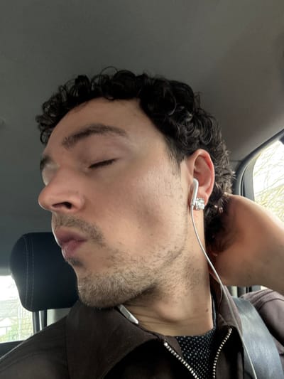 A male individual with dark, curly hair and facial stubble is depicted in a close-up, side-profile view, facing left with his eyes closed. He appears to be seated inside a vehicle, indicated by a dark headrest visible behind him and the vehicle's grey ceiling above. A white wired earphone is inserted into his left earlobe, which also features a square, reflective silver earring. His right hand is positioned behind his head, touching the back of his neck. He wears a dark brown collared jacket with a silver zipper, over a dark garment with a light polka-dot pattern. Through a vehicle window on the right side of the frame, blurred light-colored outdoor elements are visible, suggesting daylight conditions. The image does not contain any visual information to confirm the specific location as Hazel Grove, United Kingdom.