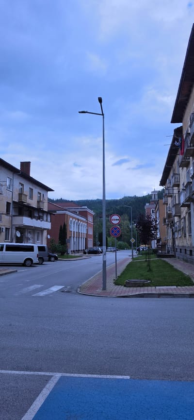 The image depicts a street scene in Rudozem, Bulgaria. The street, oriented in a receding perspective, is bordered by buildings of varying heights and architectural styles. A sidewalk is visible on the right side of the street, marked with brick paving. Several parked vehicles, including a white van, are positioned along the right side of the road. Overhead, there is a lamppost extending from the left side of the image. Further down the road, two traffic signs are visible: one indicating "No Passing" and another indicating "No Stopping." In the background, there is a green hillside. The sky is overcast with a mix of light blue and gray clouds. The overall scene suggests a residential area within the city.