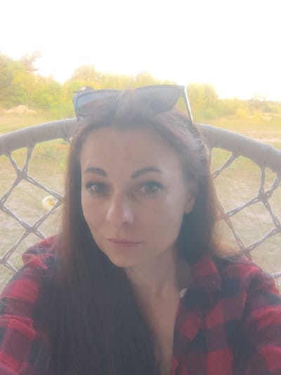 A woman with long, dark brown hair, wearing a red and black plaid shirt and sunglasses perched on her head, is seated in a hanging chair outdoors. She is looking directly at the camera with a neutral expression. The background shows a blurry landscape with trees and a bright, hazy sky, suggesting it is daytime and possibly sunset or sunrise. The location is identified as Дзівінскі сельскі Савет, Belarus.