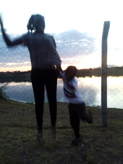 A blurry, full shot shows two figures near a fence against a lake and setting sun. The taller, silhouetted figure appears to be jumping, arm raised, while holding the hand of a smaller child who is in motion. The child wears a light shirt and dark pants. The background features calm water reflecting the sky, a thin treeline, and distant hills. The image has a grainy quality.