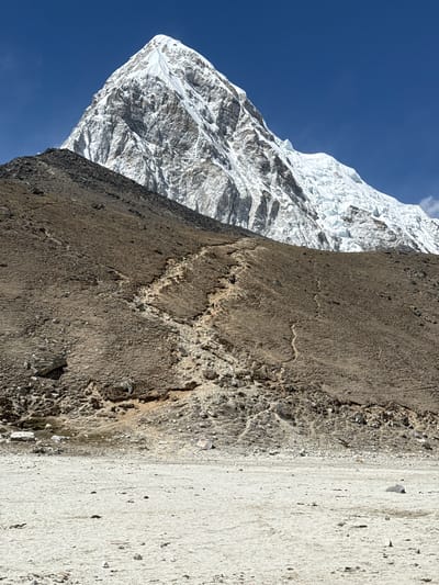 This vertically oriented image showcases a dramatic mountain landscape, likely in the Himalayas, specifically Khumjung, Nepal.
On the left side, a towering, snow-capped mountain dominates the scene, its vast expanses of white snow and ice contrasting sharply with dark, exposed rock faces. The snow appears thick and heavily textured, indicating deep glaciations and rugged terrain. The mountain rises against a clear, vibrant blue sky, suggesting excellent visibility and fine weather.
The right side of the image features a steep, rocky, and earthy foreground. This area is characterized by a coarse texture of dark brown and grey soil, loose pebbles, and irregular rock fragments. Patches of lighter, sand-colored earth are also visible, particularly towards the right edge, hinting at erosion or glacial till. The foreground's surface is uneven and rugged, emphasizing the harshness of the high-altitude environment.
The scene appears to be under bright daylight conditions, given the clear sky and well-defined shadows. There are no people, animals, or man-made objects visible, underscoring the raw, untouched nature of the landscape. No discernible activity is taking place, and no text is visible in the image.
