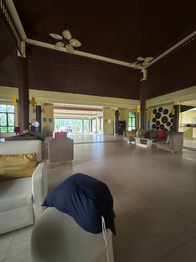 The image depicts a large, open indoor common area, possibly a resort lobby, located in Ban Bang La On, Thailand. The space features a light-colored tiled floor and light beige walls. The ceiling is high, dark brown, and pitched, supported by several dark brown wooden columns that feature golden decorative elements. Multiple light-colored, five-bladed ceiling fans are affixed to the ceiling.

In the foreground, on the left, a white armchair with a yellow cushion is partially visible. Immediately in front of it, a light-colored ottoman holds a dark blue object, possibly a bag or folded fabric, with white drawstrings visible. Further into the midground on the left, another brown armchair with a red cushion is present, and behind it, a light brown square armchair.

Towards the background, large open archways lead to an outdoor area characterized by lush green foliage and visible palm trees. Indistinct human figures are visible walking in this distant outdoor area.

On the right side of the midground, a seating arrangement includes a light brown sofa with red and yellow cushions, a coordinating light brown armchair, and a white rectangular coffee table supporting a small green plant and a green vase. Behind this seating area, a wall displays dark circular decorative elements. Further to the right background, an additional open corridor or room is visible, with what appears to be part of a balustrade or railing.

No identifiable individuals are present in the foreground or midground; indistinct human figures are visible in the distant outdoor area.