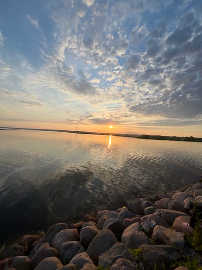 A wide-angle, eye-level shot captures a serene sunset over a calm body of water, likely the Baltic Sea, near Nasva alevik, Estonia. The sky is a canvas of blue, dotted with scattered, fluffy white clouds that are tinged with orange and yellow from the setting sun. The sun itself, a bright orb, is low on the horizon, casting a golden reflection across the water.
The water is still, with gentle ripples disturbing its glassy surface. On the far horizon, a low, green shoreline is visible, punctuated by two wind turbines standing starkly against the colorful sky. The foreground is dominated by a rocky embankment, composed of numerous large, rounded stones in varying shades of gray and brown. Some sparse green vegetation grows between the rocks. A person's foot, wearing a pink sandal, is visible on the right side of the frame, resting on the rocks, suggesting a human presence at the scene. The overall atmosphere is one of peaceful tranquility at the end of the day.