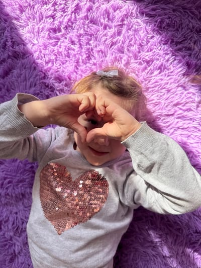 A young child lies on a purple, shag rug, wearing a grey long-sleeved shirt with a sequined heart on the chest. The child is obscuring their face with their hands, forming a makeshift 'binoculars' shape with their fingers. A small white bow is visible in the child's hair. The scene is brightly lit with natural light, creating shadows on the rug and the child. No further geographical information is discernable.