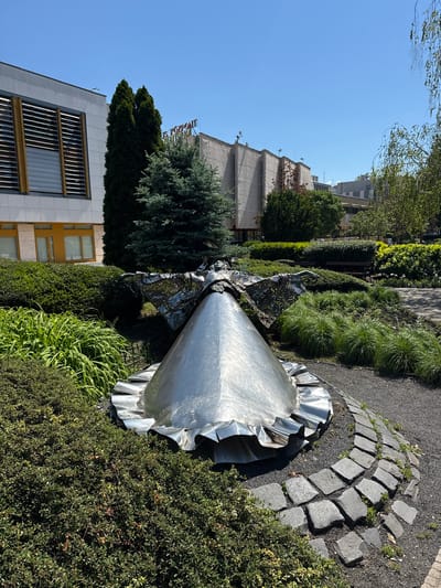 The image shows a metal sculpture in Siófok, Hungary, likely in a park or garden. The sculpture appears to be of an angel or female figure, with a flowing dress or skirt made of shiny metal. The sculpture is surrounded by green bushes and plants. A pathway of cobblestones curves around the sculpture's base. Buildings are visible in the background, including one with a restaurant sign. The sky is clear and blue. The lighting suggests it is daytime.