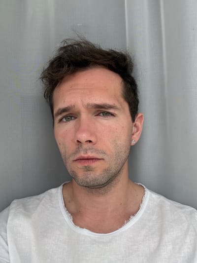 A close-up, portrait-style shot features a young man from Kurchatov, Russia, lying down and looking directly into the camera. He has dark, wavy hair, light skin with minor blemishes, and light-colored eyes, appearing green or hazel. A short stubble or light beard covers his jawline and upper lip. He wears a simple white sleeveless top, possibly made of linen, with visible frayed detailing along the neckline and armholes. A small, silver-toned, heart-shaped stud earring is visible in his left ear. The man's expression is neutral, with a hint of thoughtfulness or melancholy. The background is a plain, light grey surface, likely a sheet or curtain, suggesting an indoor setting with soft, diffused daylight. There is no visible text in the image.