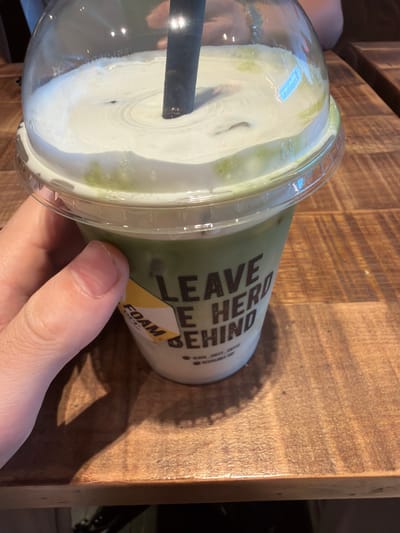 A hand holds a plastic cup containing a green-tinted beverage with a thick layer of white foam on top. A black straw is visible within the cup. The cup has a clear plastic lid and a label that reads "LEAVE HERO BEHIND" and "FOAM." The cup is positioned on a wooden surface. The image does not provide sufficient detail to identify a specific location in Bretton, United Kingdom.
