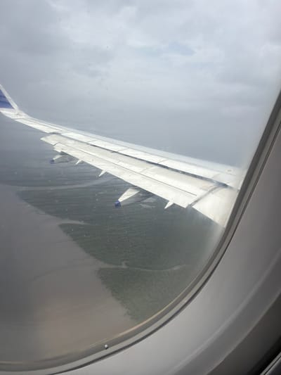 An aerial view from an airplane window shows the wing of the aircraft extended to the left. The wing is white with blue accents at the tip. Below the wing, a body of water, possibly a bay or inlet, meets land covered in green vegetation. The water appears somewhat muddy or murky. The sky above is overcast with thick gray clouds. The image is framed by the airplane's window, with the white interior of the window frame visible on the right side of the picture. The scene is likely over the coastal region near Vakrul, India, given the appearance of water and vegetation.