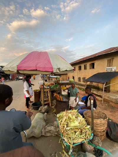 This image depicts a bustling street scene in Igarra, Nigeria, during late afternoon or early evening, characterized by soft, warm lighting. The sky above is light blue with scattered, illuminated cumulus clouds.

In the background, a large, two-story building with unpainted stucco walls and a rusty corrugated metal roof stands prominently on the right. It features multiple rectangular window openings, some with closed wooden shutters, others open. A smaller, single-story structure with a dark corrugated metal roof is attached to its front. To the far left, an unpaved road recedes into the distance, with a car and motorcycles visible.

The midground is dominated by market activity. A large, two-toned (pink and off-white) umbrella provides shade over two interconnected vendor stalls. The stall on the left displays various fruits, including watermelons, oranges, and other round items, along with bottles of liquids. A woman, wearing a white long-sleeve shirt and black leggings, stands near this stall, facing right, appearing to attend to the produce. The stall on the right features a metal drum repurposed as a charcoal grill, with several corn cobs roasting. A second woman, wearing a green t-shirt and a light-colored skirt with a headwrap, is seated on a low stool next to the grill, actively tending the corn. She is interacting with a man seated opposite her on another low stool. This man wears a white tank top, blue jeans, and a black backpack, holding a blue object and a piece of paper.

In the immediate foreground, to the left, the back of a man's head and shoulders are visible, suggesting he is seated on a motorcycle or scooter, holding handlebars. He wears a dark blue-grey long-sleeved shirt. The ground is unpaved and dusty. Various items are placed on the ground, including two large, light-colored sacks filled with unknown contents, a black bucket, a tall woven basket, and a black and red bag. A prominent pile of corn husks and unhusked corn cobs rests in a blue plastic crate on a wheeled stand in the lower right corner. A green hose runs across the lower right of the frame.