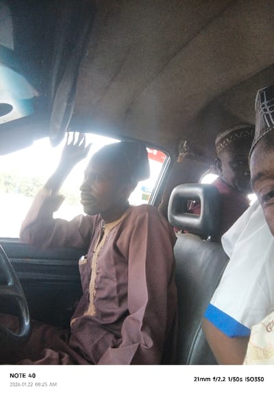 The image captures the interior of a car with three men, taken from a perspective that suggests the photographer is also inside the vehicle, possibly in the front passenger seat. The setting is identified as Kabuga, Nigeria, during daylight hours, specifically 08:25 AM according to the metadata.

In the foreground, seated on what appears to be the driver's side, is a man wearing a dark brown, long-sleeved traditional garment with light embroidery down the front, and a dark, flat-topped cap. He is looking out the side window to the left, with his mouth slightly open as if speaking or smiling. His right hand is raised towards the top of the window, partially obscured by a car visor, possibly shielding his eyes from the intense light or gesturing.

Behind him, another man is visible, looking forward. He wears a reddish-brown garment and a light-colored, intricately patterned cap. To the right, a third man's face and upper body are partially seen, wearing a white garment with a distinctive blue band on the sleeve, and a similar patterned cap.

The car's interior features a grey headliner and dark seats. A portion of the steering wheel is visible on the left side of the frame. The view outside the car windows is overexposed due to strong backlighting, indicating a bright and likely sunny morning.

Visible text in the bottom left corner includes "NOTE 40" and the capture date and time "2026.01.22 08:25 AM". In the bottom right corner, camera metadata states "21mm f/2.2 1/50s ISO350".