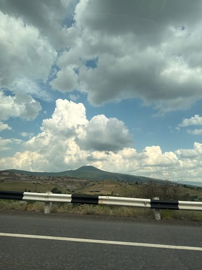 The image depicts a roadside view from the city of Arzni, Armenia. The foreground features a segment of an asphalt road with a white line marking. Adjacent to the road is a white and black striped guardrail, which appears to be constructed of metal. Beyond the guardrail, a dry, grassy embankment slopes upwards. The middle ground is characterized by rolling hills and mountainous terrain, with some areas showing signs of erosion or excavation. Scattered vegetation and a few small structures are visible on the hillsides. The sky dominates the upper portion of the image, filled with a mix of dramatic, fluffy white cumulus clouds and darker, gray storm clouds. A single, cone-shaped mountain peak is visible in the distance, silhouetted against the cloudy sky. The overall impression is a rural, mountainous landscape under a dynamic sky.