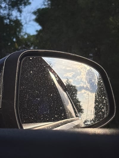 The image displays a close-up, slightly low-angle view of a car's black-framed side mirror, located in Dnp Ryzhiki, Russia. The mirror's surface is heavily encrusted with dried water spots or dirt, and a distinct vertical crack or scratch traverses the right side of the reflective glass. The mirror reflects a partly cloudy sky exhibiting white clouds and warm light, suggesting either early morning or late afternoon, above a reflection of dark green foliage. The background outside the mirror's frame consists of blurred dark green trees and sections of light blue sky. The foreground shows a dark, out-of-focus portion of the vehicle's interior, likely the dashboard or door panel. No people or discernible actions are present in the scene.
