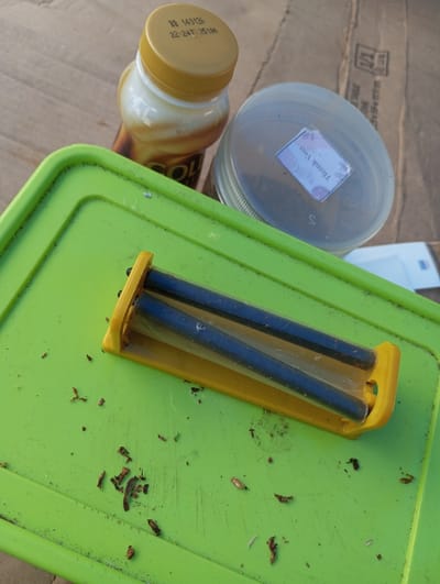 The scene depicts a collection of items arranged on a corrugated cardboard surface in Cibeureum, Indonesia. A bright green, flat, possibly plastic lid or tray occupies the central foreground. Scattered across this green surface are numerous small, dark reddish-brown fragments consistent with loose tobacco. A yellow plastic cigarette rolling machine, containing two black rollers and a transparent band, rests on the green surface towards the top right. In the background, behind the green surface, a plastic bottle with a yellow screw-top cap is visible; it contains a light brown liquid and has a label partially showing "GOLD." The cap bears the printed text "BB 140125" and "22:24 T: 2510A." To the right of the bottle, a clear plastic jar with a translucent lid is present. A small, white rectangular sticker with the text "Thank You" (oriented upside down from the primary viewing angle) and a faint floral design is affixed to the center of the jar's lid. The underlying cardboard surface is light brown and shows faint dark markings, including what appears to be part of a shipping label or box print, such as "T-11." No individuals are present.
