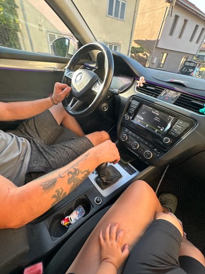 This media file shows the interior of a car, viewed from the perspective of someone sitting in the driver's seat. A person's arms are visible in the foreground; one arm has a tattoo of an arrow and feathers. The other arm, to the left, is resting on the steering wheel. A child's hand is visible reaching towards the person's leg, which is covered by a black garment. The car's dashboard features a digital display showing trip information, including time, distance, and fuel consumption. The display also shows the current temperature as 29.5 degrees Celsius. There are various buttons and controls for the car's audio system and climate control. A car key with a decorative fob is in the center console. Outside the car, through the windows, buildings and trees are visible, suggesting a sunny day in a town or village. The scene appears to be set in Oreshak, Bulgaria. The overall impression is one of a casual car journey on a warm day.
