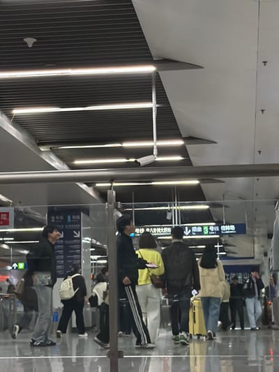 The scene depicts a concourse within Nanjing South Railway Station, located in Yuhuatai District, Nanjing, China. The environment is a brightly lit, modern indoor space with a ribbed metal ceiling, integrated linear LED lighting, and large white ceiling panels. A white surveillance camera is mounted from the ceiling, positioned to view the area below. Horizontal metal railings and vertical transparent glass panels serve as barriers. Signage in simplified Chinese and English is visible, indicating directions for "3号线 往林场" (Line 3 To Linchang) and "1号线 往八卦洲大桥南" (Line 1 To Baguazhou Bridge South).
Approximately fifteen individuals are present, all appearing to be adults, engaged in transit. In the mid-ground, a group of four people walks away from the viewer. From left to right within this group: a male in a black tracksuit with white side stripes, holding a light-colored object; a female in a bright yellow top and light-colored trousers, carrying a brown bag; a male in a dark, possibly patterned, jacket and dark trousers; and a female in a light beige fluffy jacket and blue jeans, pulling a yellow wheeled suitcase. To the far left, a male wearing a black hoodie, light blue jeans, and Adidas sneakers is positioned facing right, exhibiting an open-mouthed expression. In the background, several other individuals are visible walking or standing near what appear to be entry or exit points, one wearing a white backpack. The floor exhibits reflections of the overhead lighting and figures.