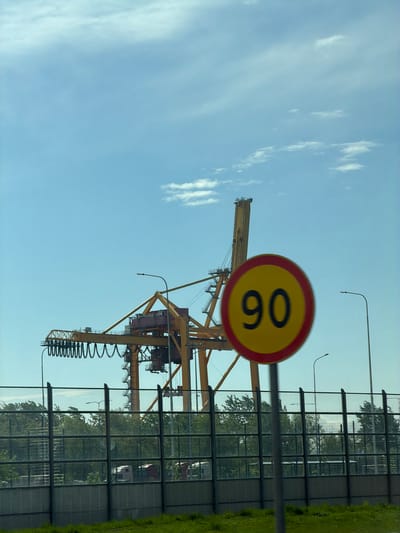 The image shows a crane in Lisy Nos, Russia, with a round speed limit sign of 60. The crane is yellow and has a visible spring mechanism. The sign is in the foreground, partially obscuring the crane. Behind the crane is a sound barrier fence, followed by trees and grass. The sky is blue with scattered clouds.