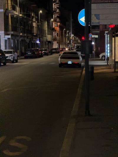 The image captures a nighttime street scene in Novara, Italy, likely on Corso Cavour, given the presence of the traffic light and the style of buildings commonly found on this major thoroughfare. A white sports car, possibly a Dodge Challenger, is parked on the right side of the street, its rear facing the camera. Its license plate is partially visible, reading "ZE 990 BG". Several other cars are parked along the street on both sides. The street is lined with multi-story buildings, illuminated by streetlights and lights from within the buildings, casting a warm glow. A blue circular traffic sign with a white arrow indicating a turn is visible on a pole to the left, along with a red traffic light. A pedestrian walkway is visible on the right, separated from the street by a curb. The overall impression is of a quiet urban night, with the city beginning to settle down.