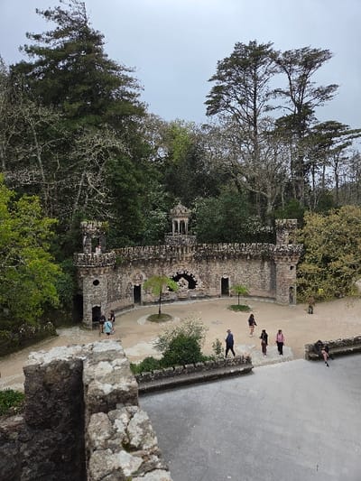 The image displays an outdoor scene at the Quinta da Regaleira estate in Sintra (Santa Maria e São Miguel, São Martinho e São Pedro de Penaferrim), Portugal, characterized by historical stone architecture and dense vegetation under an overcast sky.
In the foreground, the top of a rough-hewn stone wall, exhibiting grey and brown tones and some moss or lichen growth, partially occupies the lower left corner, indicating an elevated viewpoint.
The midground is dominated by a weathered stone wall, approximately 3-4 meters high, constructed from grey-brown rough-cut stones. This wall features two square turrets with crenellated tops on its left and right extremities. A single individual stands atop the left turret. In the center of the wall, a large, dark, arched grotto-like recess is visible. To its left and right, smaller, rectangular door-like openings provide access. Integrated stone benches are present along sections of the wall, particularly to the left of the grotto. Above the wall, slightly left of the grotto, a small, ornate, open-sided stone cupola with a dome roof is positioned.
In front of this main wall lies a light-colored, unpaved courtyard area. Two small deciduous trees with sparse foliage are centrally planted within this courtyard. Towards the right foreground of the courtyard, a darker, possibly paved, pathway or plaza area is visible.
Numerous people populate the scene. A group of three individuals, dressed in casual attire, stands or walks near the left side of the courtyard. On the right-hand paved area, four individuals are walking from left to right: one person in a dark blue jacket and hat, followed by three others in various casual jackets and pants (light top/dark skirt, dark jacket/dark pants, pink jacket/dark pants). Further to the right, a single individual is seated on a low stone wall or bench, leaning back.
The background is densely forested with a variety of mature trees, including tall conifers with visible trunks and sparse foliage against the sky, as well as deciduous trees, some displaying lighter, possibly lichen-covered, branches. The sky above is uniformly light grey and overcast.