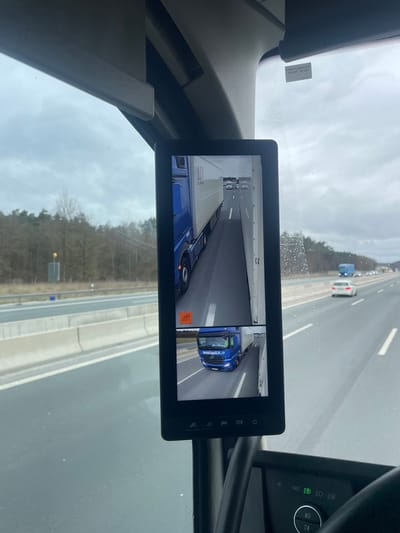 The image displays a view from inside a vehicle on a multi-lane highway, identified as being in the vicinity of Mühlhausen, Germany. The foreground features a rectangular digital screen, likely a camera monitor replacing or augmenting a mirror, mounted centrally within the vehicle's interior. This screen is split into two video feeds: the upper feed shows the side of a blue semi-truck with a white trailer, marked "AIT," moving alongside other vehicles on the highway. An orange warning icon is present in the lower-left of this feed. The lower feed shows the front-left perspective of a different blue Mercedes-Benz semi-truck, also with a white trailer, positioned on the same highway.
Outside the vehicle, through the windshield, an overcast sky is visible above a multi-lane asphalt highway that appears damp. Concrete barriers line the median. Scattered vehicles, including a white sedan and another blue truck, are present on the highway, indicating traffic flow. Bare trees and vegetation line both sides of the road in the middle distance. Portions of the viewing vehicle's interior, including the ceiling, upper dashboard, and a steering column with illuminated light controls, are visible at the periphery of the frame. No individuals are discernible in the scene.