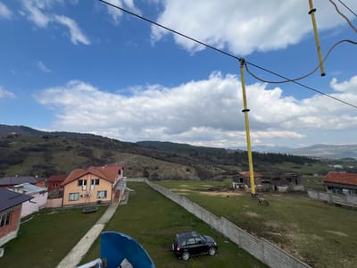 The image depicts an elevated view of a semi-rural landscape in Rakitovo, Bulgaria, under a partially cloudy blue sky with scattered white cumulus clouds.

In the foreground, a partial blue structure is visible at the lower left. A black compact SUV is parked on a manicured green lawn, adjacent to a narrow concrete pathway that leads towards an orange two-story house with a red-tiled roof, white-framed windows, and two external air conditioning units on its side. A trampoline is situated on the lawn next to this house. Several other residential buildings with varied roof colors are partially visible to the left and behind the orange house.

A long, segmented gray concrete block fence runs diagonally from the mid-left to the mid-right, separating the closer residential area from a more expansive green field with patches of bare earth. Beyond this fence, two horses (one dark gray, one light brown) are grazing in the field. Further to the right, several rustic wooden barn-like structures with dark roofs are present. Indistinct human figures are visible near one of these wooden structures, and a dark bird is on the ground nearby.

Overhead, black utility cables extend horizontally across the upper portion of the frame, supported by yellow vertical poles. One prominent yellow pole extends downwards from a cable near the center-right, and a curved brown cable is also visible in this overhead network.

The background consists of rolling hills and mountains, sparsely vegetated with grass, bushes, and patches of forest, extending towards a distant mountain range under the cloudy sky.