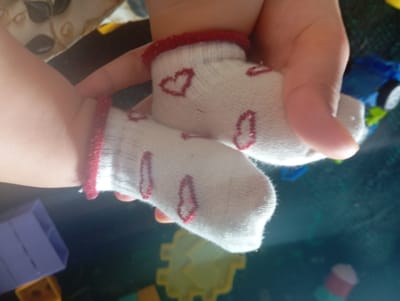 The image shows a close-up, high-angle view of a child's feet wearing white socks with red heart patterns. The socks have red ribbed cuffs. A person's hands are visible, holding the child's feet. In the background, out of focus, are some children's toys, including a blue toy with wheels and purple building blocks. The scene appears to be indoors on a dark, reflective surface. The location of Kızıl-Bel, Kyrgyzstan is not discernible from the image itself.