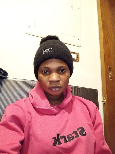 A female individual is depicted in a chest-up portrait within an indoor setting in Madinaty, Egypt. She is wearing a black knitted beanie hat with a pom-pom and white "NYC" embroidery on the front, along with a bright pink hooded sweatshirt featuring a cowl neck. Black, reversed text, possibly "Stark," is visible on the front of the sweatshirt. A small stud piercing is present on her right nostril. The background consists of a light-colored wall, displaying a white electrical panel and a small, ornate, gold-colored rectangular object. To the subject's left is a grey, vertically slatted structure, potentially a radiator. A wooden door with a brass doorknob is partially visible to her right.