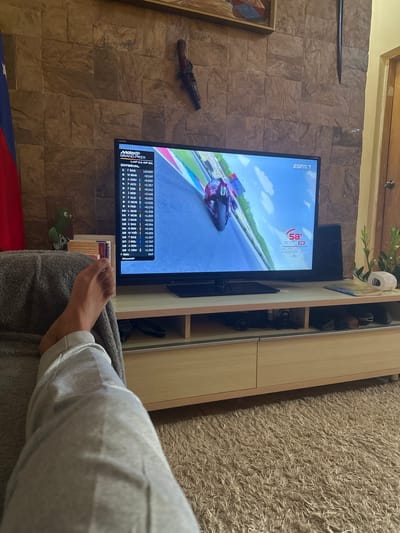 The image captures a living room scene where a person is watching a motorcycle race on a television. The television screen displays a MotoGP Grand Prix, with rider standings and lap information visible. The room has a rustic aesthetic, with a stone-tiled accent wall behind the television. To the left of the TV, a decorative antique-style gun is mounted on the wall. To the right of the TV, a light-colored wooden entertainment unit is visible, with shelves containing various items, including what appear to be speakers and possibly cameras. A plush, light brown carpet covers the floor, and a person's leg, clad in grey sweatpants, is in the foreground, suggesting they are comfortably seated or reclining. A blanket is draped over their lap. The overall impression is a relaxed, indoor setting, likely in the evening or during daylight hours given the artificial lighting of the room. The text on the screen indicates a racing event, with rider names, lap times, and positions. The text "ESPN 7" suggests the broadcast channel. A small green stuffed toy is visible near the bottom left of the frame.