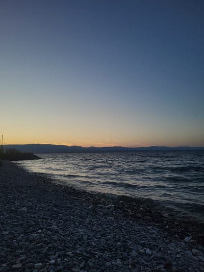 A tranquil seascape is depicted with the sky on the left transitioning from blue to orange near the horizon. To the right, water with gentle waves touches a rocky shoreline. The pebbles on the shore are dark and closely packed. The scene is simple, focusing on the elements of nature.