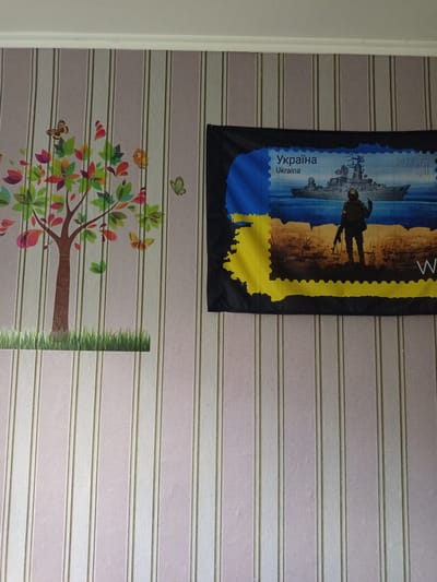 The image shows a section of a wall in a room. The wall is covered with striped wallpaper, predominantly in light pink and white, with thin vertical lines of a slightly darker shade. On the left side of the wall, there is a colorful sticker of a tree with various butterflies and leaves. The tree has a brown trunk and branches, and the leaves are in shades of green, pink, red, and yellow. Several butterflies, depicted in brown, orange, and yellow, are scattered around the tree. To the right of the tree, on the upper portion of the wall, a small green butterfly sticker is visible.
Dominating the right side of the image is a dark fabric, likely a flag or banner, featuring a postage stamp design. The stamp depicts a military scene with a warship at sea and a soldier standing on land, holding a rifle and pointing with one hand. The stamp incorporates the colors of the Ukrainian flag, blue and yellow, with the text "Україна" and "Ukraina" written in the upper left corner. The year "2022" is also visible in the upper right corner of the stamp. The word "W" in large white font is partially visible on the dark fabric below the stamp. The scene appears to be indoors, with a white ceiling or molding visible at the top of the image. No specific location in Pavlohrad, Ukraine can be identified from the visual information provided.
