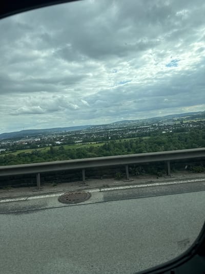 A panoramic view of a city landscape is captured from a high vantage point, likely a bridge or elevated road, looking out over Weitersburg, Germany. The foreground features a gray asphalt road with a white line marking its edge. To the left, a circular metal manhole cover is embedded in the pavement. Beyond the road, a metal guardrail runs horizontally across the image, supported by visible posts. This guardrail separates the road from a grassy embankment with some sparse vegetation.
The middle ground is dominated by a dense, green forest that stretches across the landscape. Interspersed within and beyond the forest are numerous buildings, indicating a populated area, with a river or body of water visible in the distance. The background shows a sprawling city with a mix of residential and industrial structures, extending towards a range of hills under a cloudy, overcast sky. The clouds are thick and gray, with some lighter patches suggesting breaks in the cloud cover, and a faint blue sky is visible in some areas. The overall impression is one of a slightly muted, possibly humid, day with a wide view of urban and natural scenery.