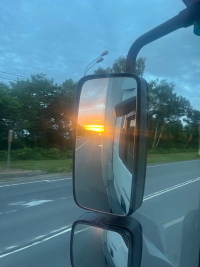 The image is a reflection in a truck's side mirror, showing a road with a sunset. The sky is cloudy and bluish-gray. Trees line the side of the road. The sun is low in the sky, casting a bright orange glow. The road has two lanes with white markings. The truck's white body is visible to the right of the mirror. The location is not specified to be in the city of Газопроводск, Russia.
