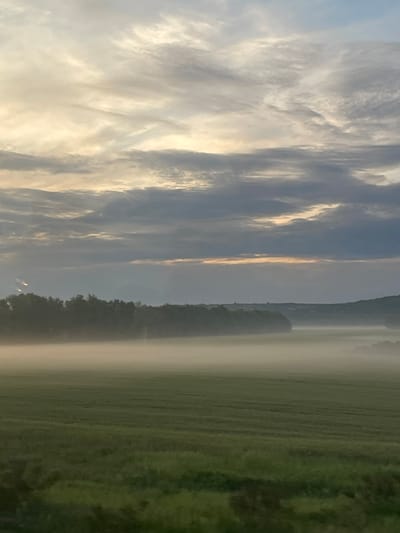 The image shows a landscape with a field covered in mist or fog. A line of trees separates the field from the sky. Above the trees, the sky is filled with clouds. The image is likely taken in a rural area near Obrenovac, Serbia.