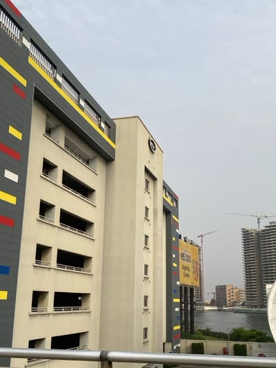The image presents an urban vista in Falomo, Lagos, Nigeria, under an overcast sky. A metallic railing extends horizontally across the lower portion of the frame. The central focus is a multi-story, light-cream colored building, likely a parking garage, featuring several open levels with visible railings. Its left side is clad in dark grey paneling accentuated with irregular horizontal stripes in red, yellow, and white. A dark-skinned individual's head and shoulders are visible, protruding from an upper-level opening on the left side of this structure.

To the immediate right, a taller, cream-colored building displays a circular 'MTN' logo near its upper edge and has a vertical arrangement of small rectangular windows. Further right, a prominent yellow billboard advertises "N5,000 Bigwhiz Combo" in black text and is affixed to a support structure with matching striped paneling.

In the background, a body of water, consistent with a lagoon or creek, separates the midground from a distant urban area. Across the water, multiple construction cranes punctuate the skyline above several partially completed high-rise buildings with exposed concrete frameworks. The sky is uniformly overcast.