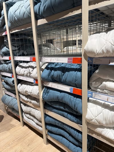 The image depicts a shelf unit in a retail setting, possibly a department store in Ragusa, Italy. The shelves contain stacks of bedding products, including quilts and comforters, displayed in various colors: white, blue, and gray. Each item is packaged with a tag indicating its dimensions, price, and the "BASIC" brand name. Labels include descriptions of product names and material composition, indicating information such as "Occasione" and "STORFJELLET," along with the size of the item (e.g., "240 x 220 cm"). The shelves are made of light-colored wood and wire, creating a grid-like structure. The floor is wood-colored. There are no people visible.
