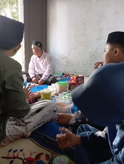 The image depicts an indoor scene in Bulukandang, Indonesia, featuring three individuals seated on a patterned blue mat overlaying a floral rug.
On the far left, partially obscured, is a person facing away from the viewer, wearing a dark green long-sleeved shirt and a black peci. They are seated cross-legged, wearing white pants with black patterns.
In the mid-ground, an older man with grey hair and a black peci adorned with a gold emblem is seated cross-legged, wearing a light pink long-sleeved shirt and dark patterned sarong or pants. He is looking towards an open window or doorway on the left, through which daylight and some foliage are visible. His hands are clasped in front of him.
On the right, another person is seated with their back partially to the viewer, wearing a dark blue top (possibly a hijab or hooded garment) and a black peci. Their bare feet are visible, and their right hand holds a thin white string or thread, suggesting an activity. They wear blue pants.
Between the individuals, an array of objects rests on the mat. These include a red rectangular box with gold designs, several transparent and opaque plastic containers (some green, some yellow), a clear plastic container filled with light brown granular material, and multiple small clear glass cups. The background features a plain, light-colored wall with a textured finish.