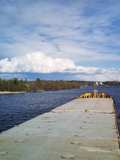 The image depicts a view from the deck of a barge on a waterway, possibly the Volga-Baltic Waterway, near Анхимово, Russia. The barge's deck is grey and has numerous circular access hatches. The stern of the barge has yellow-colored structures. The waterway is flanked by wooded shorelines on either side. A lock system is visible further down the waterway. The sky is partly cloudy, with some large cumulus clouds. High-voltage transmission towers are also visible.