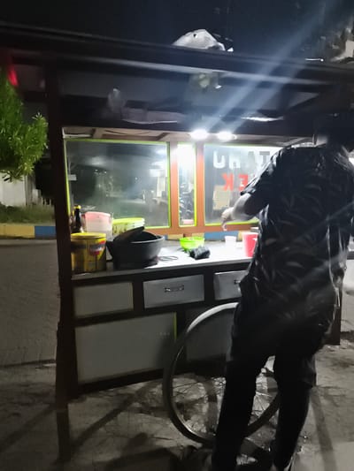 The image depicts a food cart at night on a street in Sidorejo, Indonesia. A person, viewed from behind, is standing at the cart. The cart has a dark frame with glass panels and illuminated signage that reads "CHAT EK". On the counter of the cart, there are various items including containers, a mortar and pestle, and cups. A large bicycle wheel is attached to the side of the cart. The ground appears wet and reflective, suggesting it may have rained recently. The background shows a dimly lit street with some vegetation and buildings.