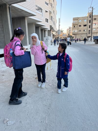This image depicts a street scene in Mu'adammiyat ash-Sham, Syria, during daytime with clear skies.
In the foreground, three young females, likely school-aged children, are standing on a dirt and gravel path adjacent to a paved road. The girl on the left, with dark hair in a long braid adorned with a purple and white flower clip, wears a pink long-sleeved shirt, dark cargo pants, and black athletic shoes. She carries a pink backpack containing a visible water bottle and a dark blue tote bag. She faces slightly towards the center. The central girl wears a white hijab, a pink long-sleeved button-up shirt, dark blue jeans, and white athletic shoes, carrying a yellow backpack. Her right hand is near her mouth, and she looks directly towards the viewer. The girl on the right, the youngest, has dark hair in two braids and wears a dark blue long-sleeved tunic-style uniform with pink bow embroidery and trim, dark blue pants, and white and pink athletic shoes. She carries a pink backpack and her left hand is touching the central girl's yellow backpack, while she looks towards the central girl.
Behind the girls, partially constructed or renovated multi-story buildings with light-colored brick facades and exposed concrete are visible on the left. Stacked cinder blocks are present near the building base. On the right, older, weathered multi-story residential buildings line the street.
The paved road extends into the background, where a group of approximately 15-20 individuals, many appearing to be schoolchildren with backpacks, are walking away from the camera. Several adult males are also interspersed within the group. Utility poles with overhead wires and a street sign are visible along the road. A blue truck is partially visible on the far right. The overall light suggests morning or late afternoon.