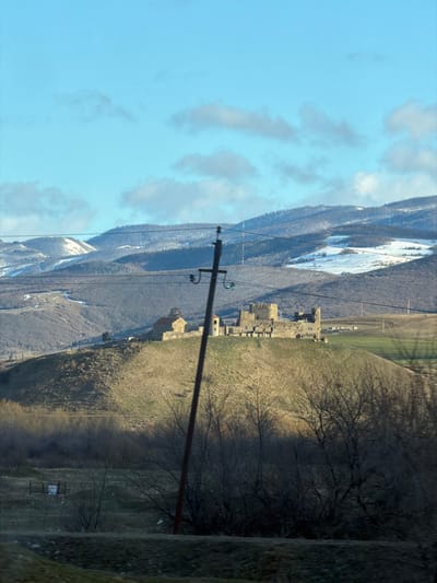 An outdoor wide shot depicts a mountainous landscape in Akhalsopeli, Georgia, under a clear blue sky with scattered clouds. In the foreground, a dark utility pole with cross arms and wires rises from the lower-center, slightly angled to the right, obscuring a portion of the scene. Below the pole, the ground is dark and uneven, with bare deciduous trees and shrubs. To the far left, a small section of a wire fence with a white sign is partially visible.
The midground is dominated by a sunlit, grassy hill with dry, golden-brown vegetation. Perched atop this hill are two stone structures: to the left, a smaller building with a reddish-brown domed roof and stone walls, consistent with a church or chapel; to its right, a larger, partially ruined stone fortress or castle with multiple towers and wall sections. Green patches of grass are visible immediately surrounding these structures, particularly on the right side of the hill. A faint track runs along the right slope of the hill.
In the background, a series of undulating hills and mountains extend horizontally across the frame. Many of these higher elevations are partially covered with patches of snow. The lower slopes of the distant mountains are covered with a mix of bare deciduous trees and dark evergreen conifers. Several thin, dark lines, likely power lines or cables, traverse the upper background horizontally. The scene is illuminated by natural sunlight, creating distinct shadows on the midground hill, indicating a low sun angle. No individuals are visible.
