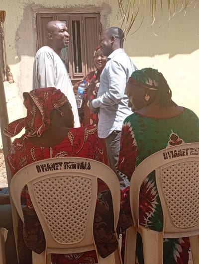 The image captures an outdoor scene in Unguwar Gaya, Nigeria, under direct sunlight. In the foreground, two individuals are seated facing away from the viewer on white plastic chairs, each bearing the visible black text "BYYANET RENTALS" on its backrest. The person on the left wears a red and black patterned dress and headwrap, while the person on the right wears a green, red, and white patterned dress and a green headwrap, with a gold-colored heart-shaped earring visible on their right ear.
In the midground, three individuals are standing and interacting. On the left, a man with a shaved head and short beard wears a light-colored, long-sleeved traditional garment; his mouth is slightly open, suggesting speech. To his right, a woman in a dark patterned dress and head covering smiles while holding a clear plastic water bottle in her right hand. To her right, a second man with short dark hair, wearing a light blue long-sleeved collared shirt, smiles and faces towards the first man. The three appear to be engaged in a positive conversation.
The background consists of a plain, light-colored wall with a rectangular window featuring multiple panes and a dark frame. Partial green foliage, possibly palm fronds, is visible in the upper right quadrant of the image. The ground appears to be unpaved dirt.