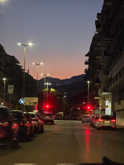 The image depicts a street scene in Salerno, Italy, at dusk. A line of cars is stopped at a red traffic light. Streetlights illuminate the road and buildings. In the background, hills are visible against the twilight sky. The architecture suggests an urban environment, with multi-story buildings lining the street. The overall impression is one of a quiet evening in a city.