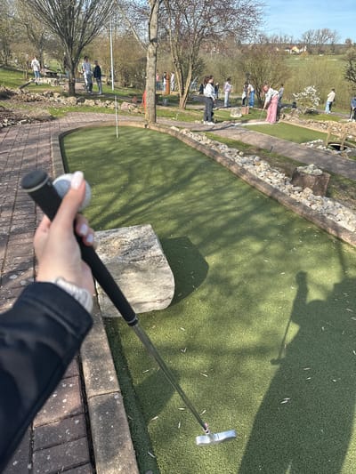 The photograph shows a person holding a putter on a mini golf course. The course has green artificial turf and is lined with rocks and concrete edging. In the background, other people are playing mini golf. The location is potentially in Gunzenhausen, Germany. The image suggests an outdoor setting with trees and some buildings in the distance. The photo is taken from the first-person perspective of someone about to putt.